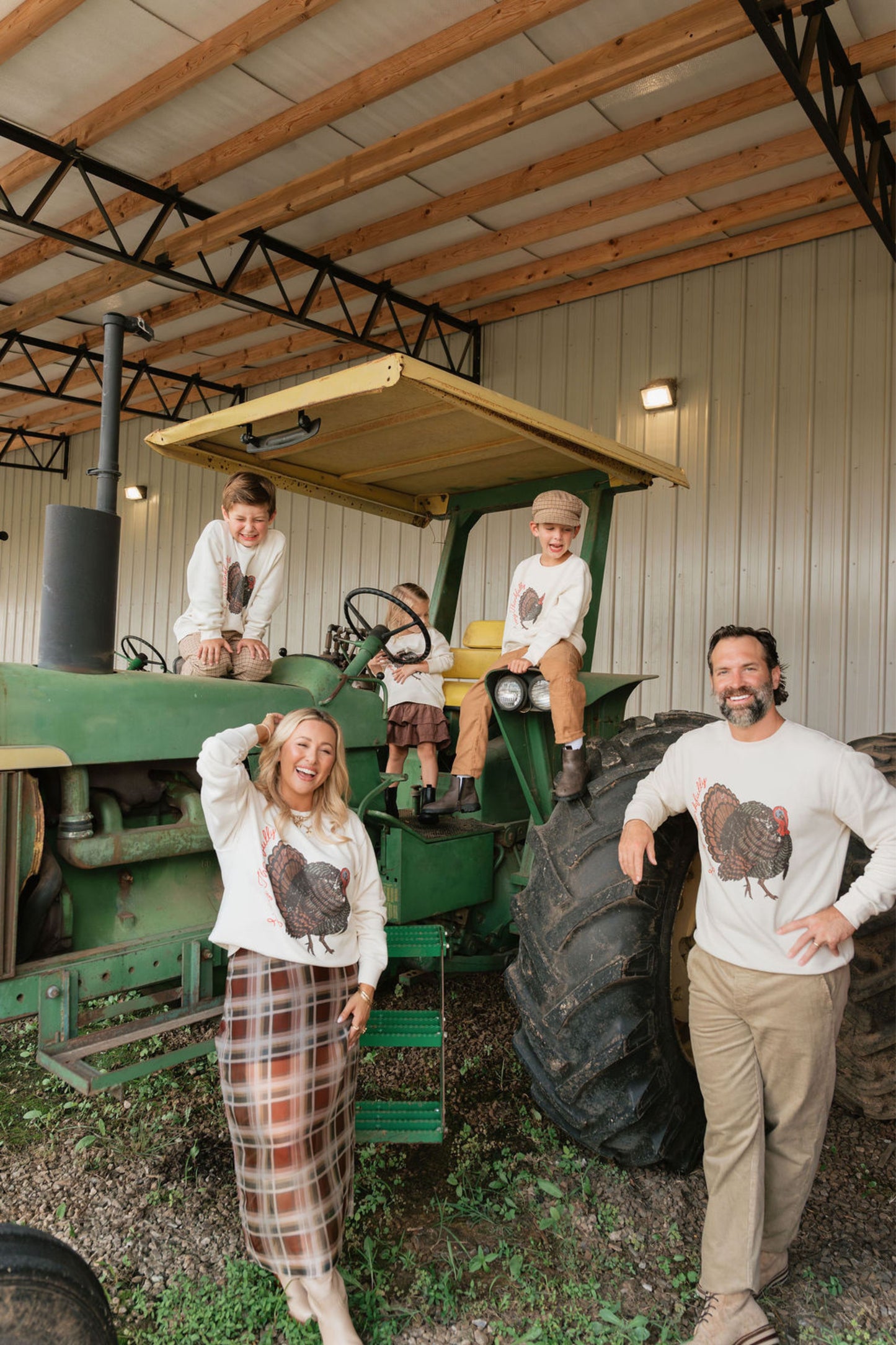 A family of five posing in a barn with a vintage green tractor. They are wearing matching white long-sleeve t-shirts featuring a graphic of a brown rooster, with the woman wearing a plaid skirt and the man wearing khaki pants.