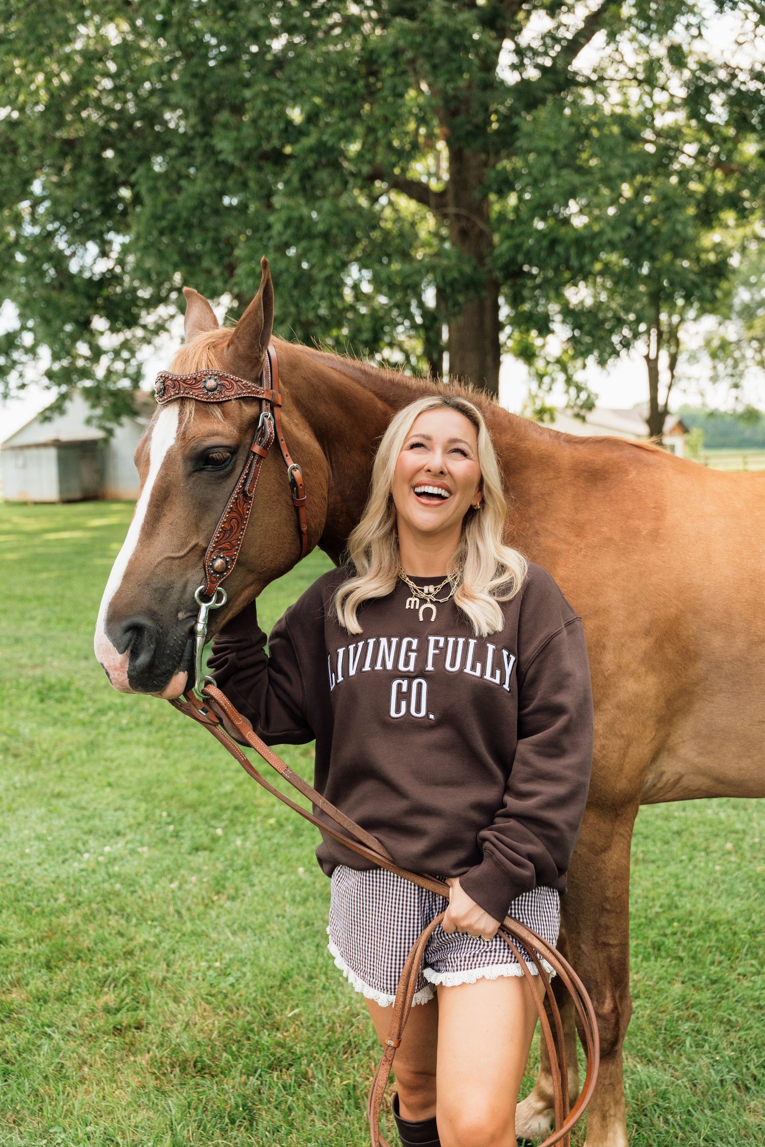 A woman smiling outdoors next to a brown horse, wearing a dark brown sweatshirt with 'LIVING FULLY CO.' printed in white across the chest.