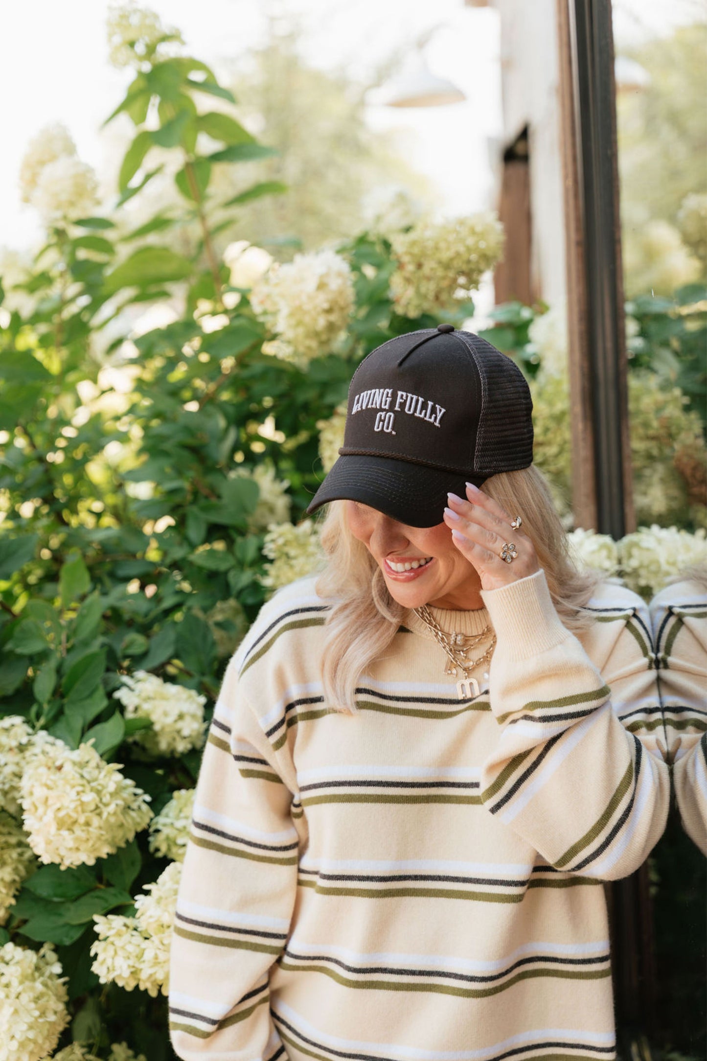 A woman wearing a black trucker hat with 'LIVING FULLY CO.' embroidered in white on the front, paired with a cream and olive striped sweater, posing outdoors in front of white hydrangea flowers.
