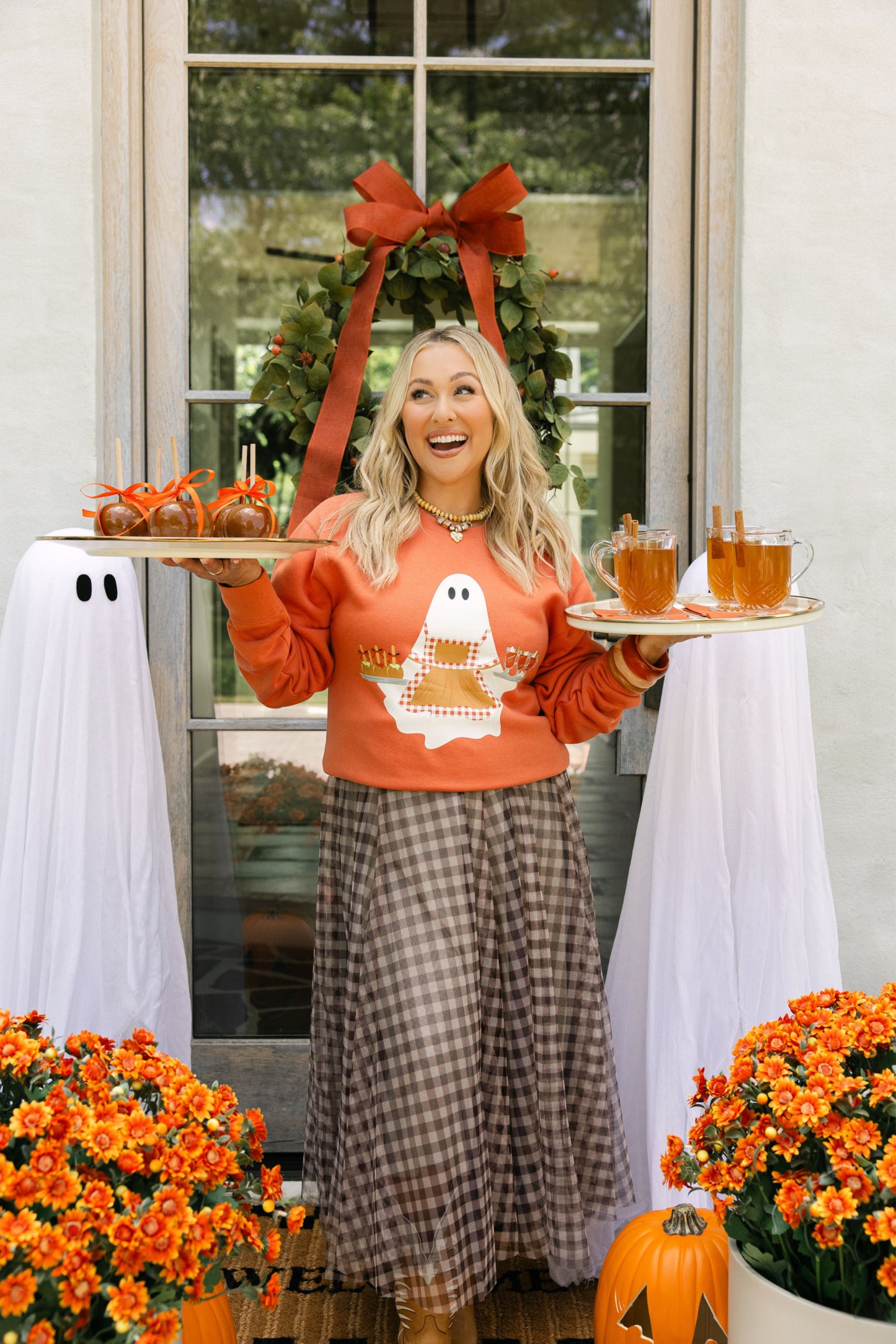 Woman in an orange sweatshirt with ghost design holding drinks, standing in front of a decorated door with pumpkins and flowers.