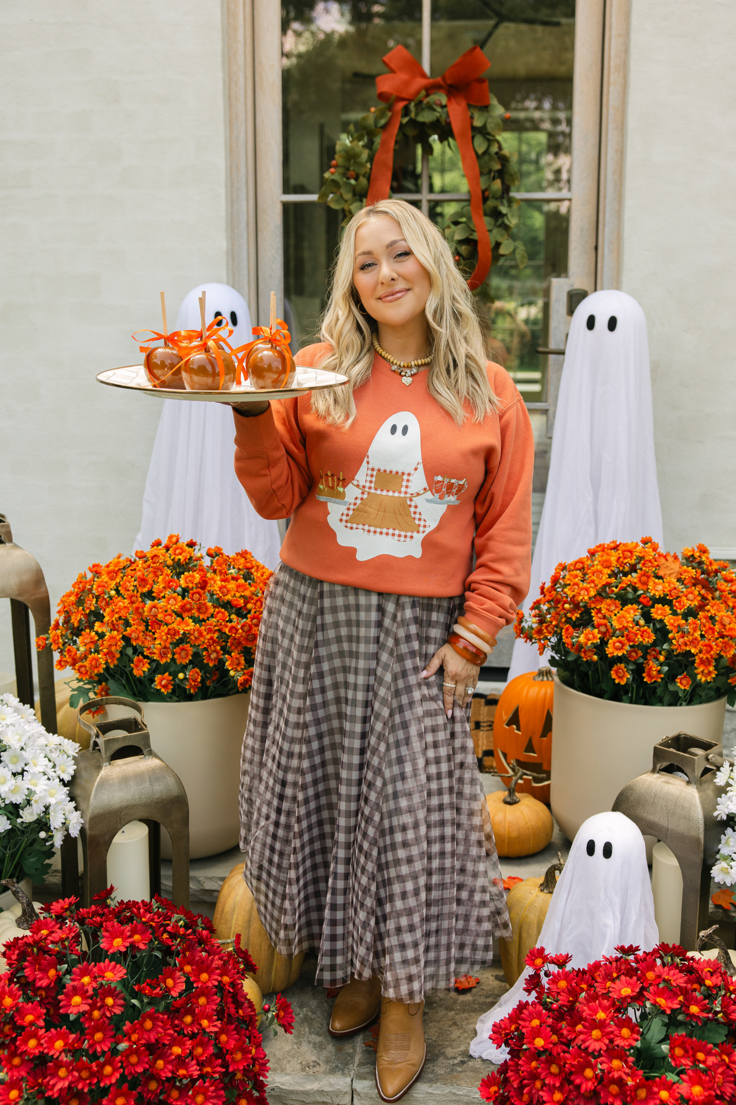 Woman standing on front porch in an orange sweatshirt with a ghost on the front holding a tray of caramel apples.
