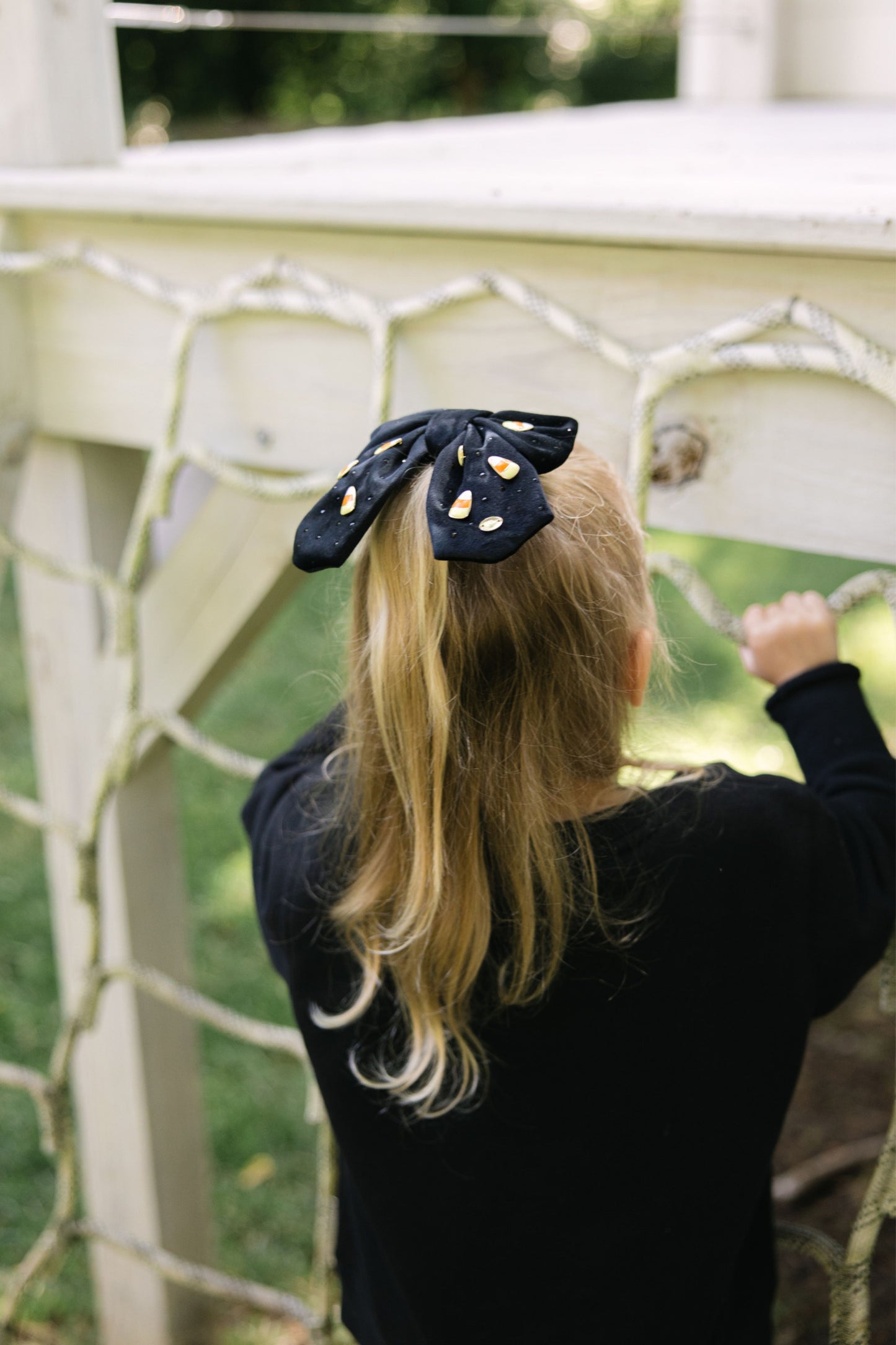 Child wearing a black hair bow with yellow and orange candy corn gems, standing in front of a white lattice structure.
