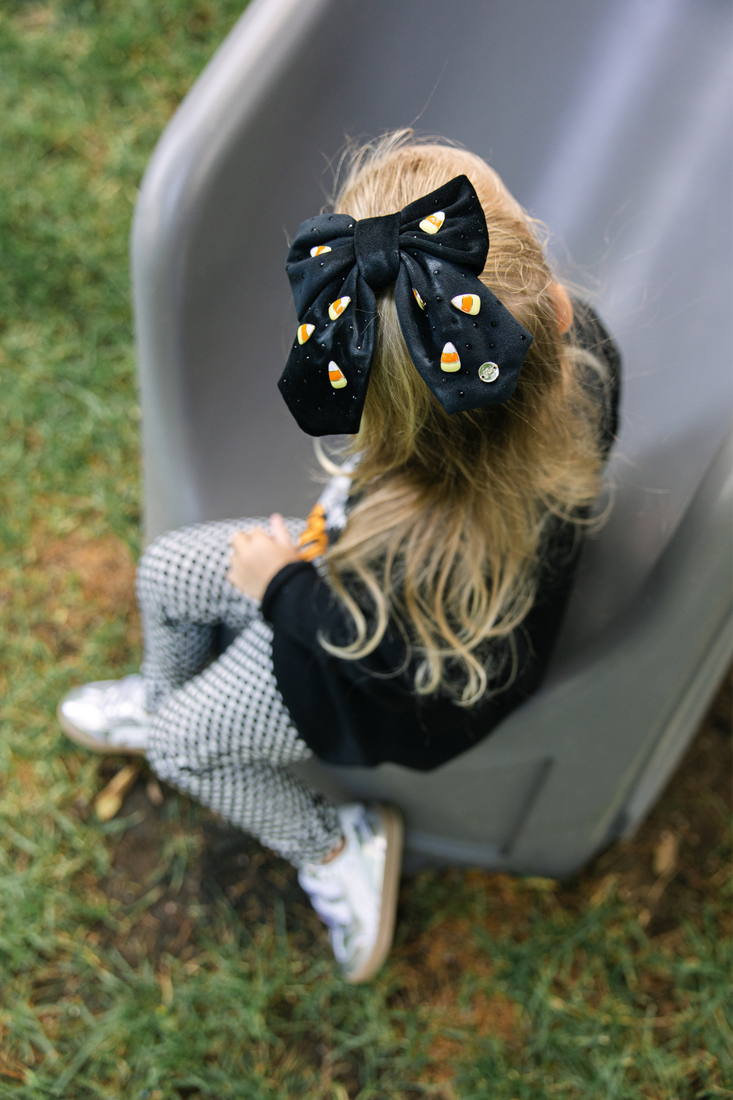 Child wearing a black bow with candy corn patterns, sitting on a gray slide outdoors.
