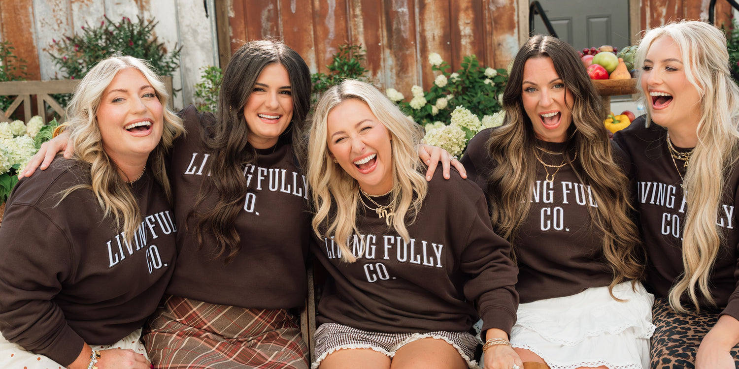 Five women laughing together while wearing matching dark brown crewneck sweatshirts with white text that reads 'LIVING FULLY CO.' in a rustic outdoor setting.
