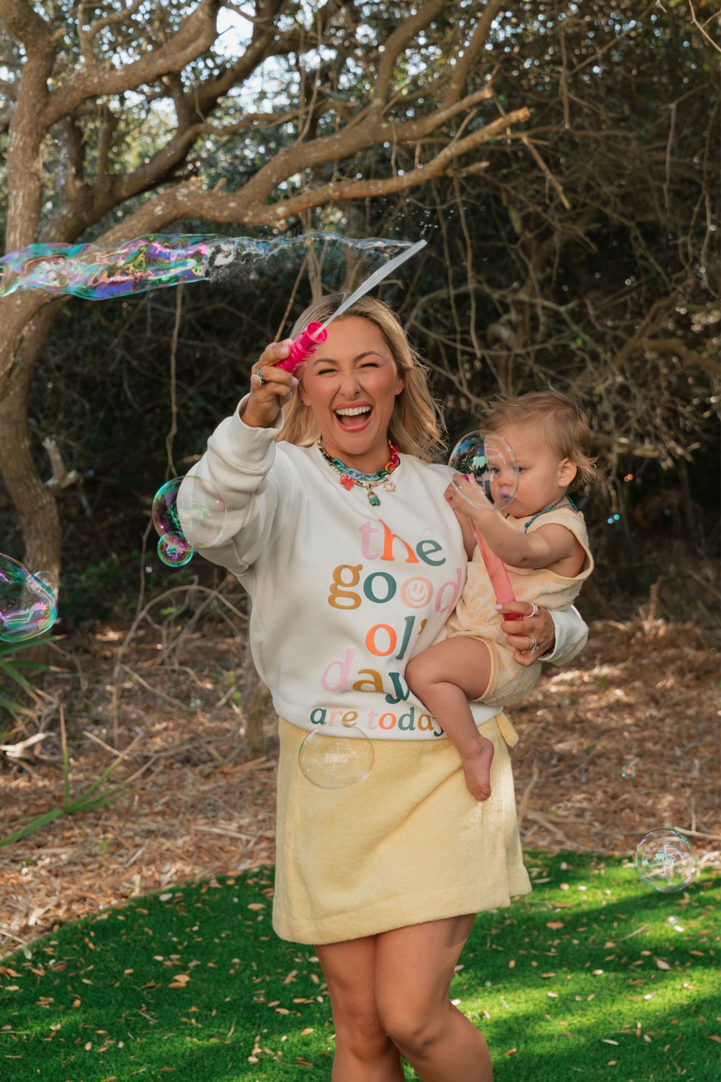 Woman holding a baby outdoors with trees and grass in the background.