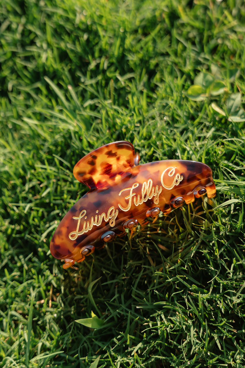 A tortoiseshell hair claw clip with gold cursive lettering that reads 'Living Fully Co', resting on a green grass background.