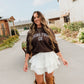 A woman walking outdoors on a paved path wearing a dark brown long-sleeve crewneck sweatshirt with 'LIVING FULLY CO.' printed in white across the chest, a white tiered ruffled mini skirt, and tall brown suede boots.