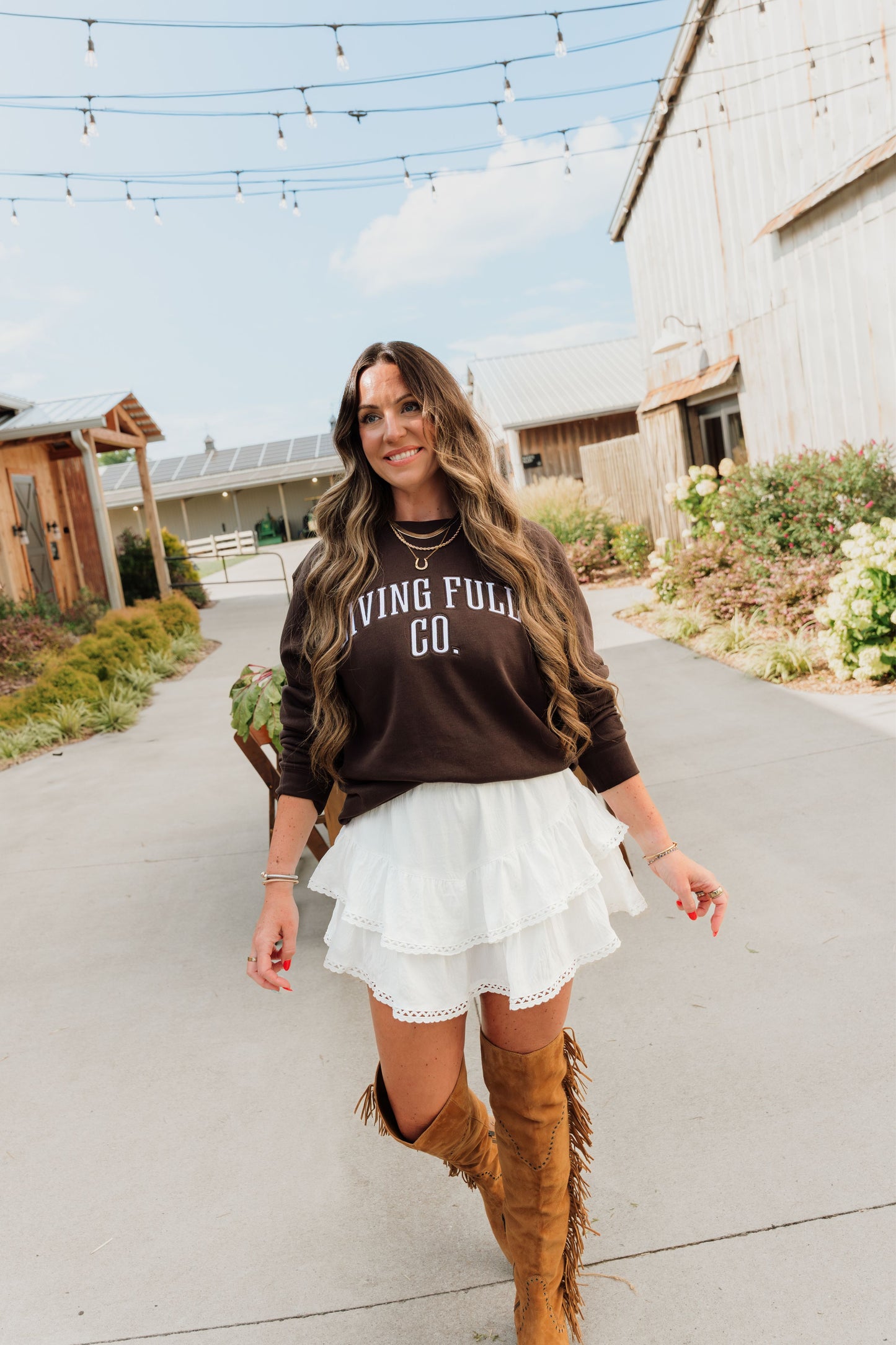 A woman walking outdoors on a paved path wearing a dark brown long-sleeve crewneck sweatshirt with 'LIVING FULLY CO.' printed in white across the chest, a white tiered ruffled mini skirt, and tall brown suede boots.