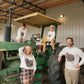 A family of five posing in a barn with a vintage green tractor. They are wearing matching white long-sleeve t-shirts featuring a graphic of a brown rooster, with the woman wearing a plaid skirt and the man wearing khaki pants.