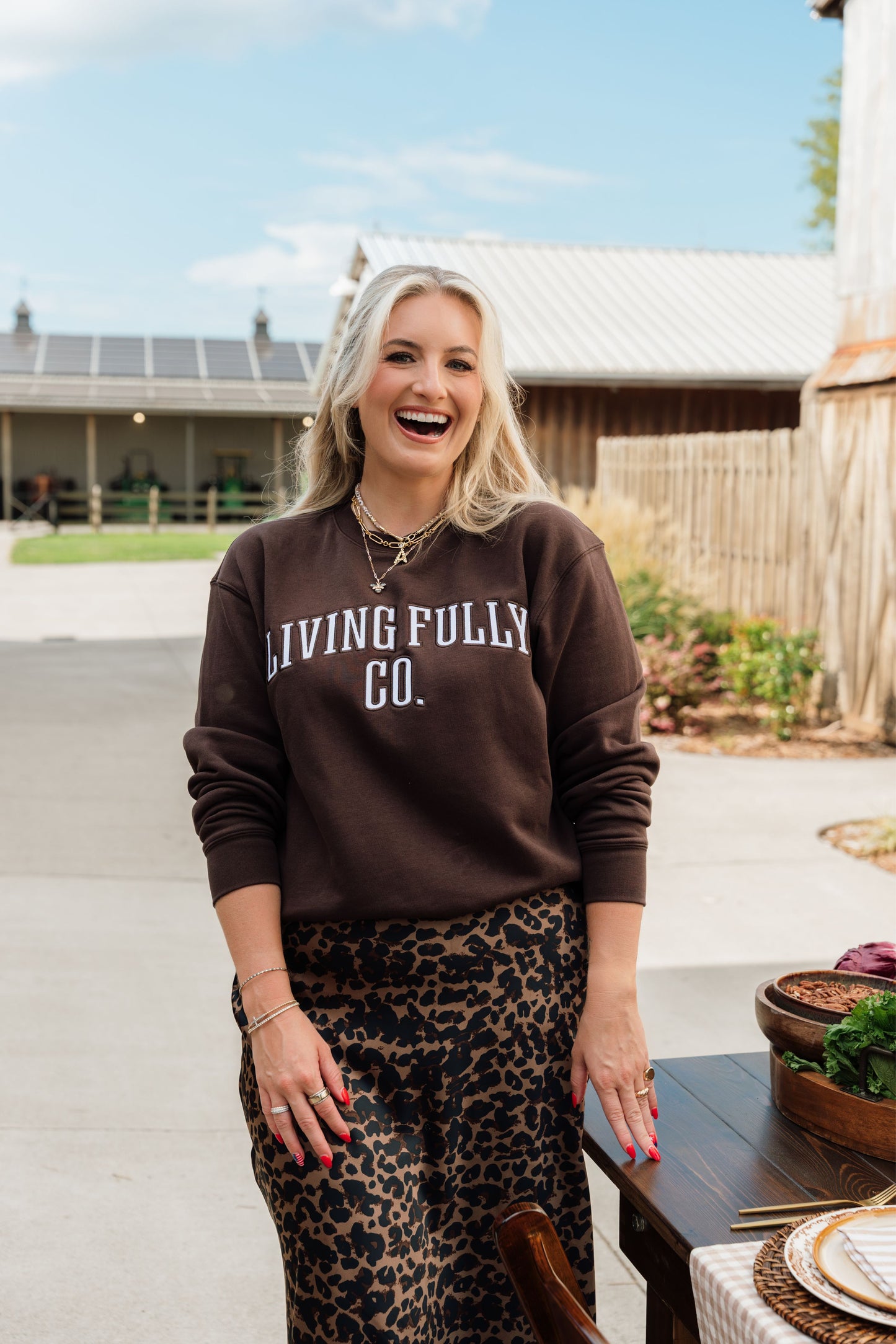 A woman stands outdoors wearing a dark brown sweatshirt with 'LIVING FULLY CO.' printed in white across the chest, paired with a leopard-print skirt.