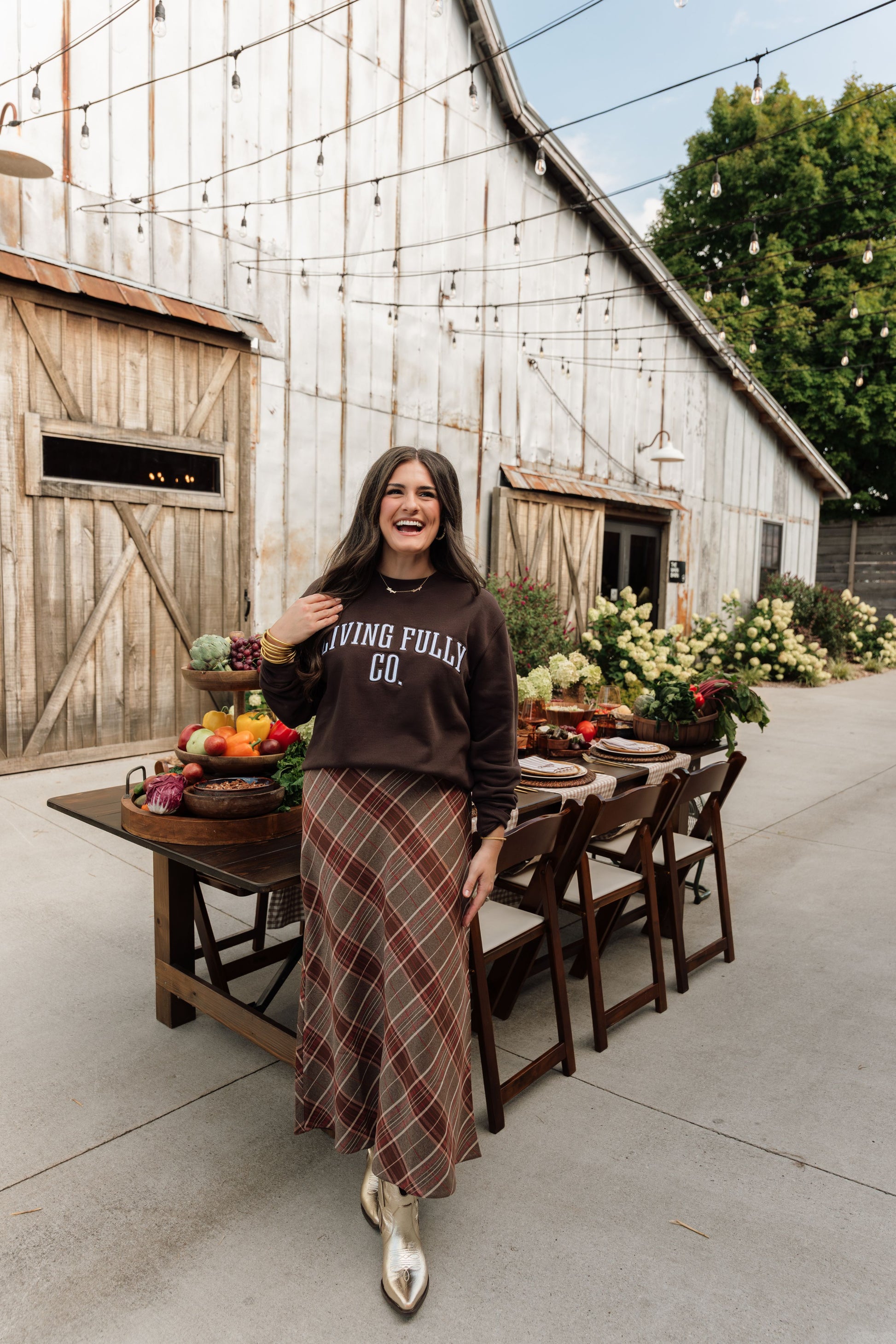 A woman stands outdoors in front of a rustic barn, wearing a dark brown sweatshirt with 'LIVING FULLY GO.' printed in white across the chest and a long brown plaid maxi skirt.