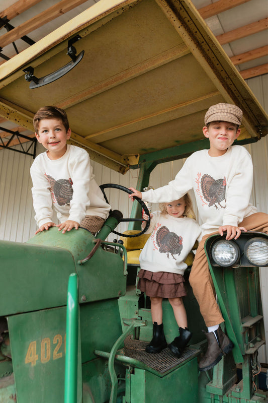 Three children wearing matching cream long-sleeve shirts with a brown turkey graphic, posed on a vintage green tractor inside a barn.