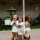 Three children standing under a covered barn with tractors in the background, wearing matching white sweatshirts with a turkey graphic and light-colored pants.
