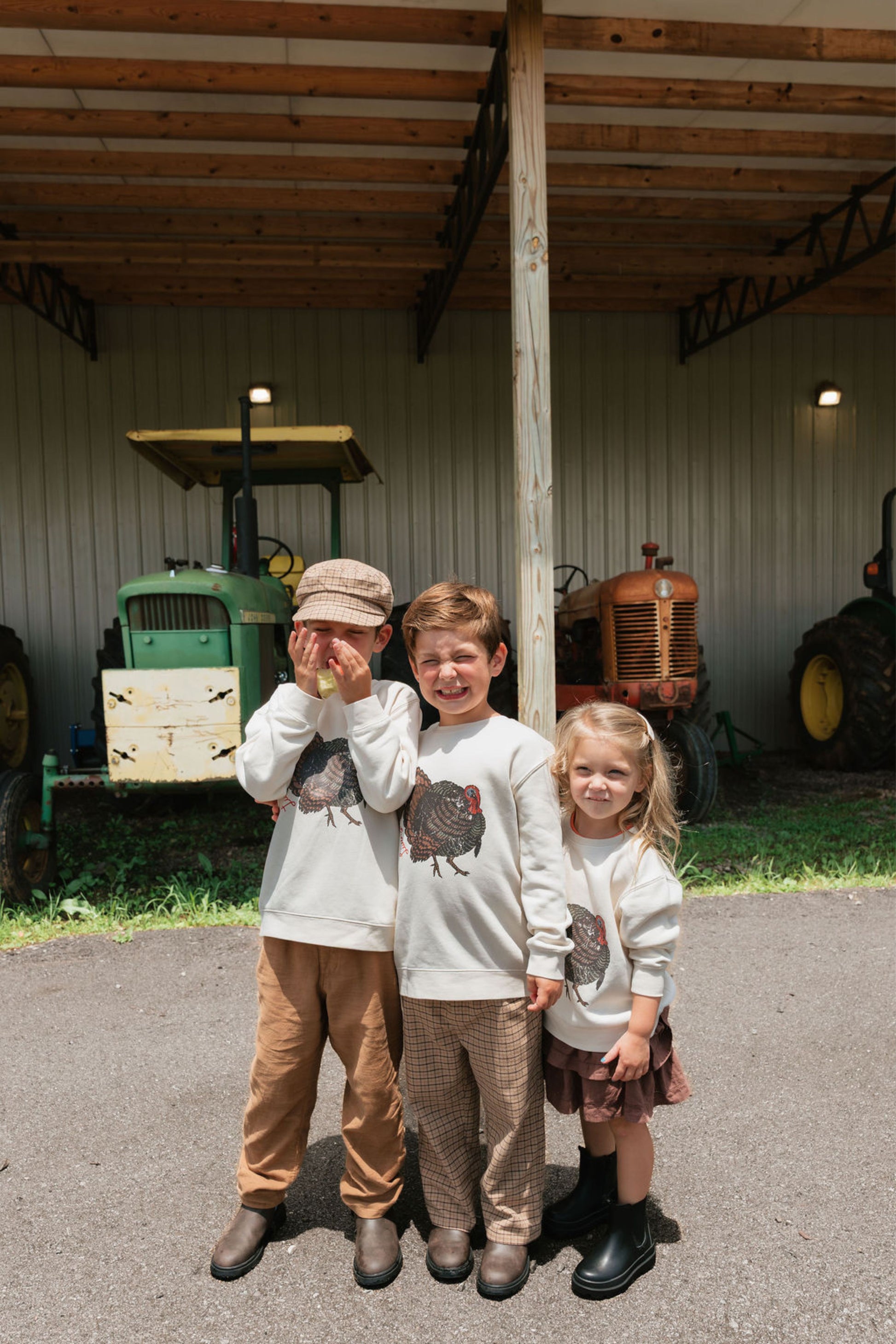Three children standing under a covered barn with tractors in the background, wearing matching white sweatshirts with a turkey graphic and light-colored pants.