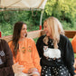 Two women laughing and sitting together on a vehicle under a canopy, wearing graphic sweatshirts. One sweatshirt is bright orange with pumpkin motifs, and the other is black with a white llama graphic and text.
