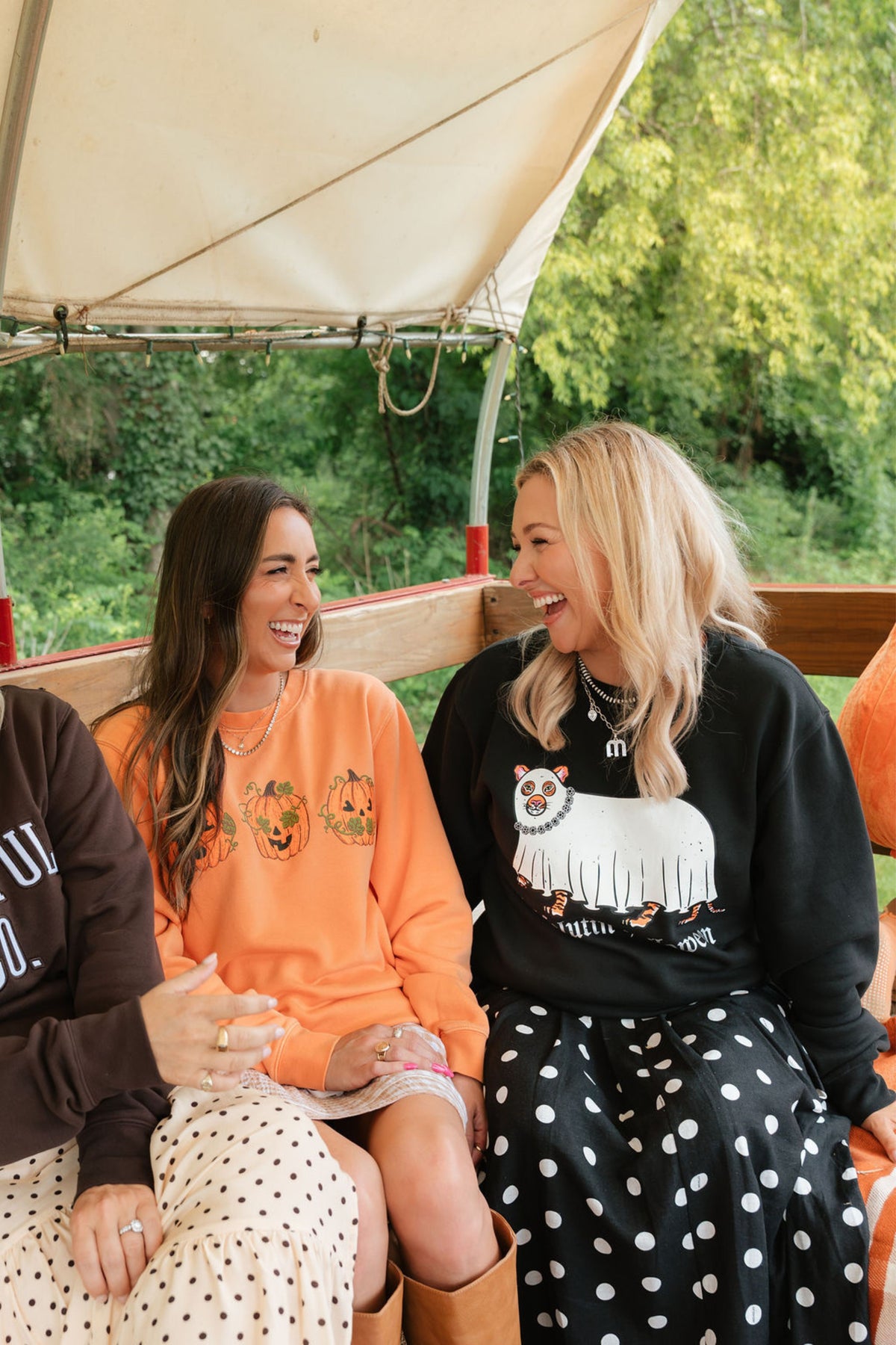 Two women laughing and sitting together on a vehicle under a canopy, wearing graphic sweatshirts. One sweatshirt is bright orange with pumpkin motifs, and the other is black with a white llama graphic and text.