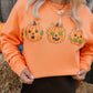 A woman wearing a bright orange crewneck sweatshirt with three embroidered jack-o'-lantern pumpkins across the chest, paired with a black skirt in an outdoor hay bale setting.