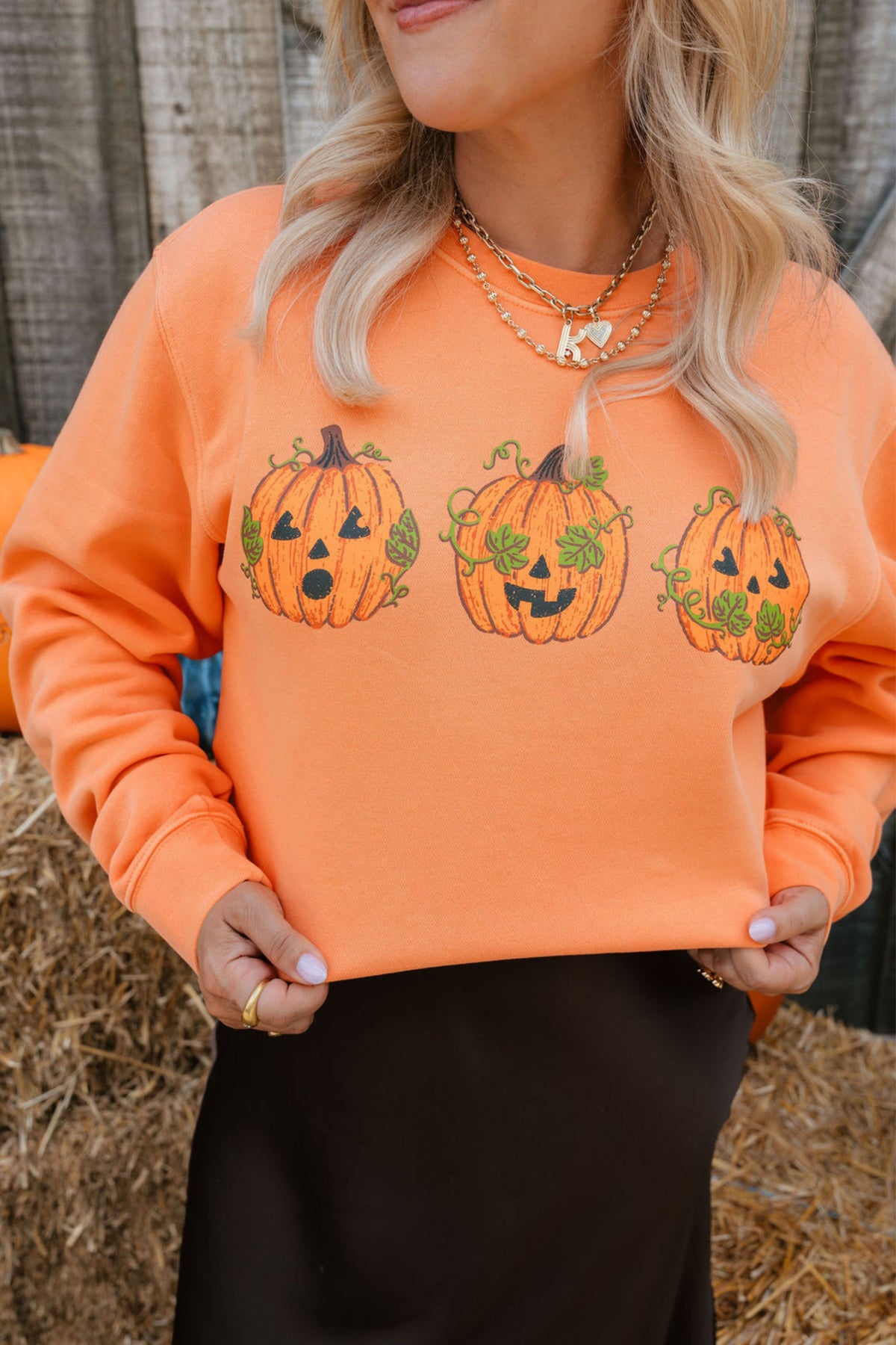A woman wearing a bright orange crewneck sweatshirt with three embroidered jack-o'-lantern pumpkins across the chest, paired with a black skirt in an outdoor hay bale setting.