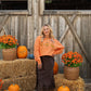 A woman stands in front of a rustic wooden barn door surrounded by pumpkins, hay bales, and orange flowers, wearing an orange crewneck sweatshirt with pumpkin graphics and a dark brown skirt.