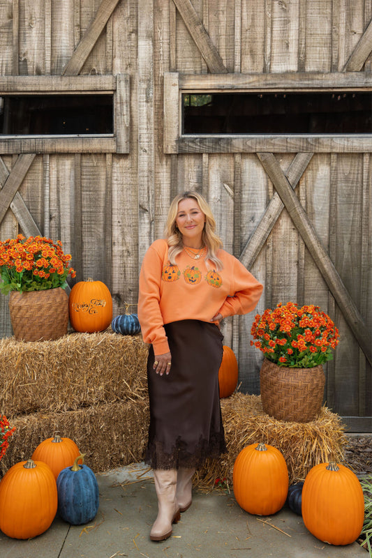 A woman stands in front of a rustic wooden barn door surrounded by pumpkins, hay bales, and orange flowers, wearing an orange crewneck sweatshirt with pumpkin graphics and a dark brown skirt.