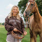 A woman smiling outdoors next to a brown horse, wearing a dark brown sweatshirt with 'LIVING FULLY CO.' printed in white across the chest, holding a coiled leather rope.