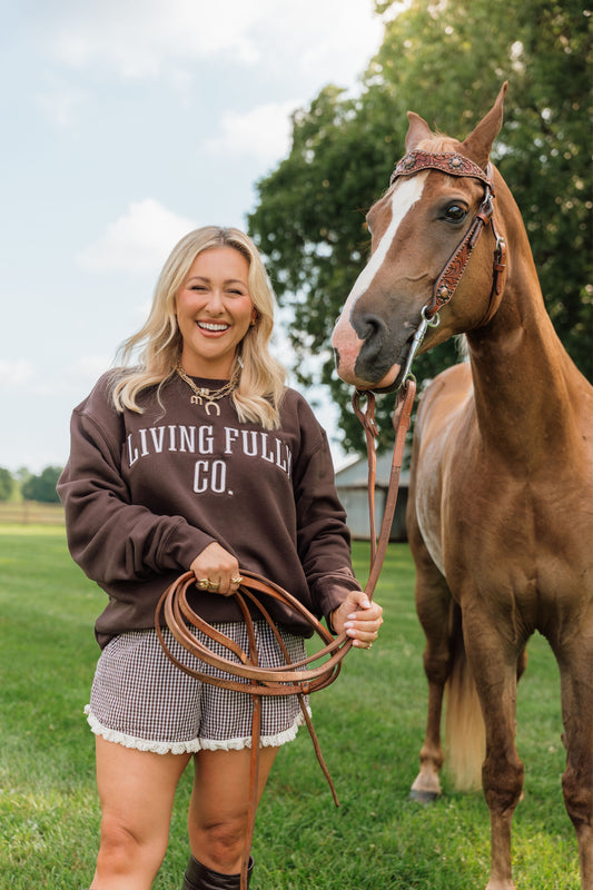 A woman smiling outdoors next to a brown horse, wearing a dark brown sweatshirt with 'LIVING FULLY CO.' printed in white across the chest, holding a coiled leather rope.