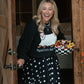 A woman stands in an open rustic wooden doorway holding a bowl of colorful candy. She is wearing a black sweatshirt with a white graphic and a long black skirt with white polka dots.