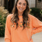 A woman smiling outdoors while wearing a bright orange crewneck sweatshirt with a pumpkin graphic across the chest, paired with a plaid skirt.