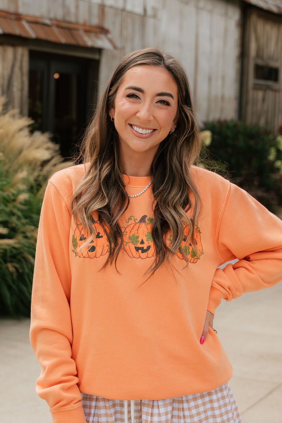 A woman smiling outdoors while wearing a bright orange crewneck sweatshirt with a pumpkin graphic across the chest, paired with a plaid skirt.