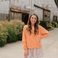 A woman stands outdoors in front of a rustic wooden barn, wearing a peach-orange crewneck sweatshirt with floral embroidery on the chest, a pink and white gingham mini skirt, and tan knee-high boots.