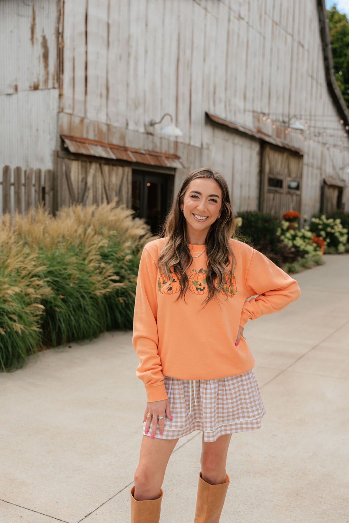 A woman stands outdoors in front of a rustic wooden barn, wearing a peach-orange crewneck sweatshirt with floral embroidery on the chest, a pink and white gingham mini skirt, and tan knee-high boots.