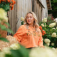 A woman with blonde hair wearing a bright orange sweatshirt with a pumpkin graphic, standing in a garden setting in front of a weathered wooden barn wall.