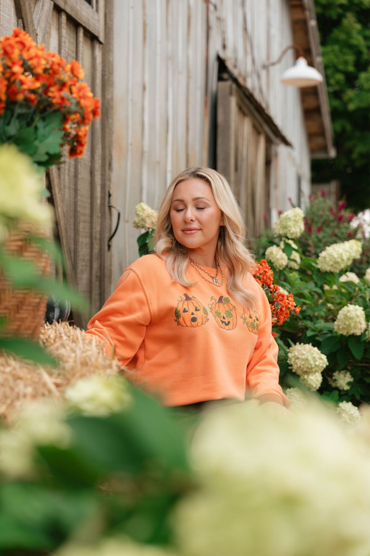 A woman with blonde hair wearing a bright orange sweatshirt with a pumpkin graphic, standing in a garden setting in front of a weathered wooden barn wall.