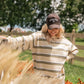 A woman outdoors wearing a cream and olive green striped knit sweater and a dark brown baseball cap with 'LIVING FULLY CO' embroidered on the front. She is smiling and posing in a garden with tall grass in the foreground.