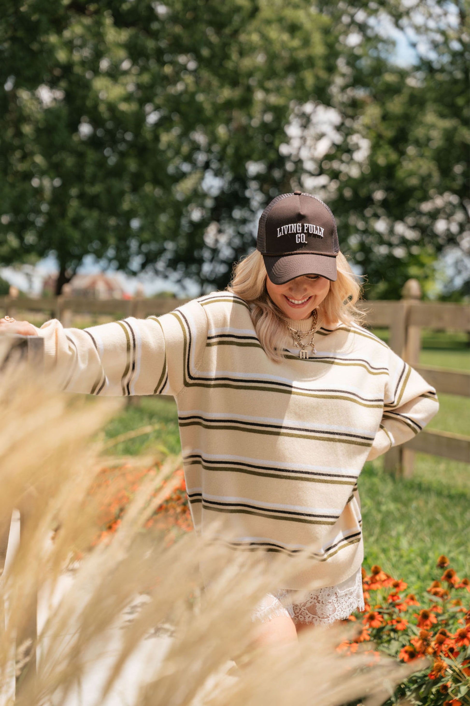 A woman outdoors wearing a cream and olive green striped knit sweater and a dark brown baseball cap with 'LIVING FULLY CO' embroidered on the front. She is smiling and posing in a garden with tall grass in the foreground.