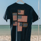 navy t-shirt with American flag designs on a hanger against a beach background