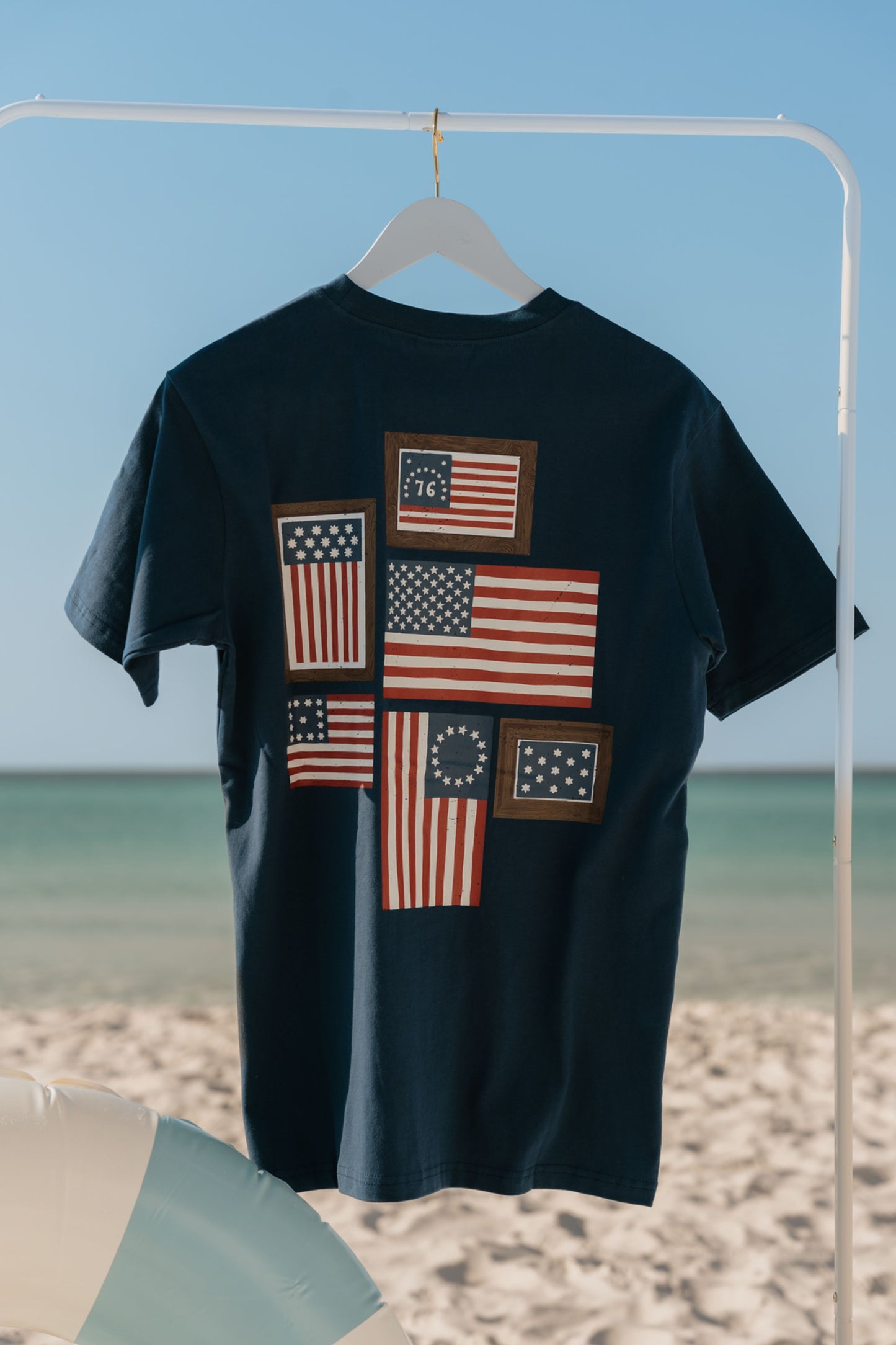 navy t-shirt with American flag designs on a hanger against a beach background