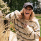 A woman outdoors wearing a cream and olive green striped long-sleeve sweater, a dark gray baseball cap with 'Living FULLY CO' embroidered on it, and smiling broadly.