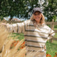 A woman wearing a beige and olive green striped long-sleeve sweater and a dark baseball cap stands outdoors in a grassy field with flowers and trees in the background.