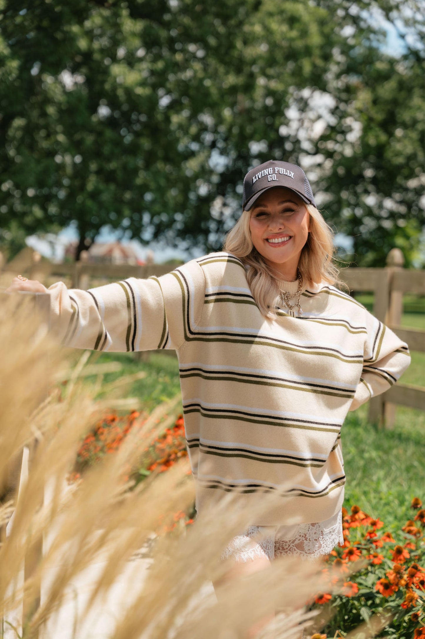 A woman wearing a beige and olive green striped long-sleeve sweater and a dark baseball cap stands outdoors in a grassy field with flowers and trees in the background.