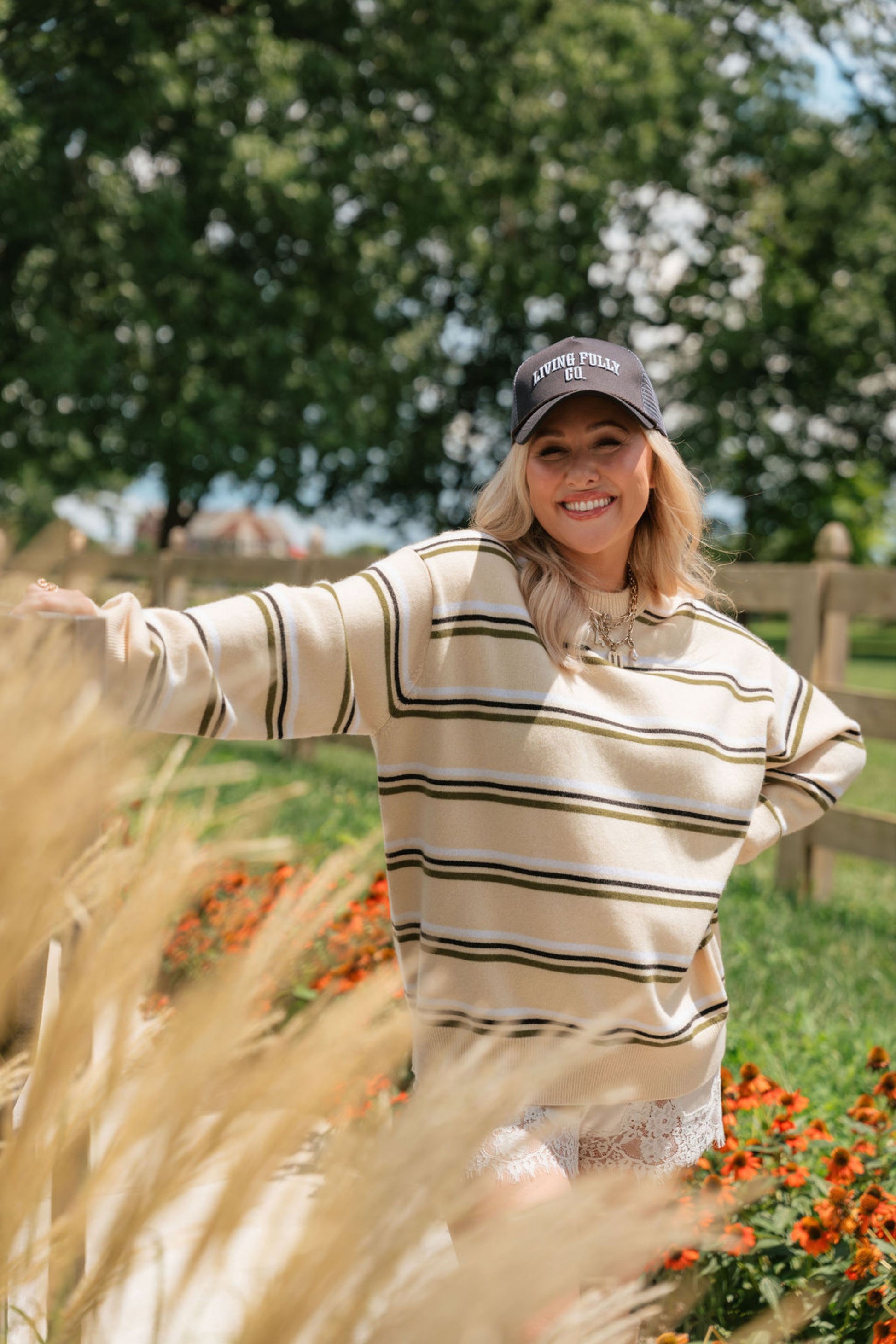 A woman wearing a beige and olive green striped long-sleeve sweater and a dark baseball cap stands outdoors in a grassy field with flowers and trees in the background.