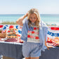 Woman in a blue and white striped outfit standing by a table with food and a patriotic tablecloth by the beach.