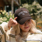 A woman outdoors wearing a dark brown trucker hat with 'LIVING FULLY CO.' embroidered in white on the front, paired with a cream and olive striped sweater.