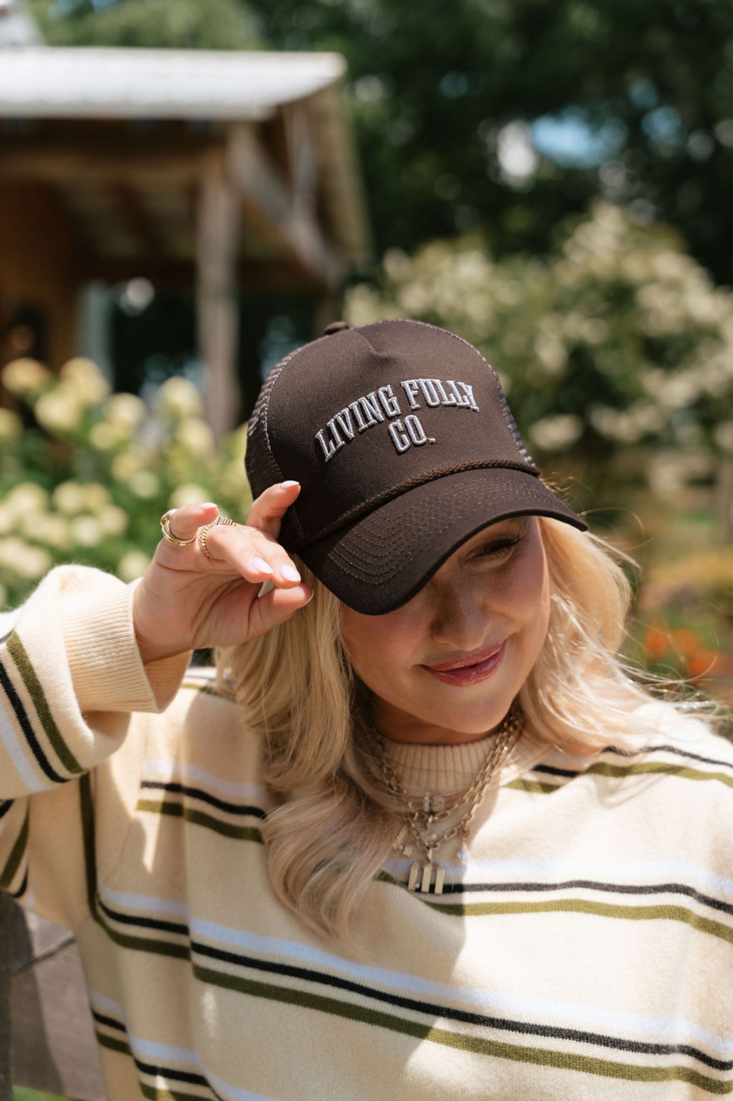 A woman outdoors wearing a dark brown trucker hat with 'LIVING FULLY CO.' embroidered in white on the front, paired with a cream and olive striped sweater.