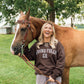 A woman smiling outdoors next to a brown horse, wearing a dark brown sweatshirt with 'LIVING FULLY CO.' printed in white across the chest.