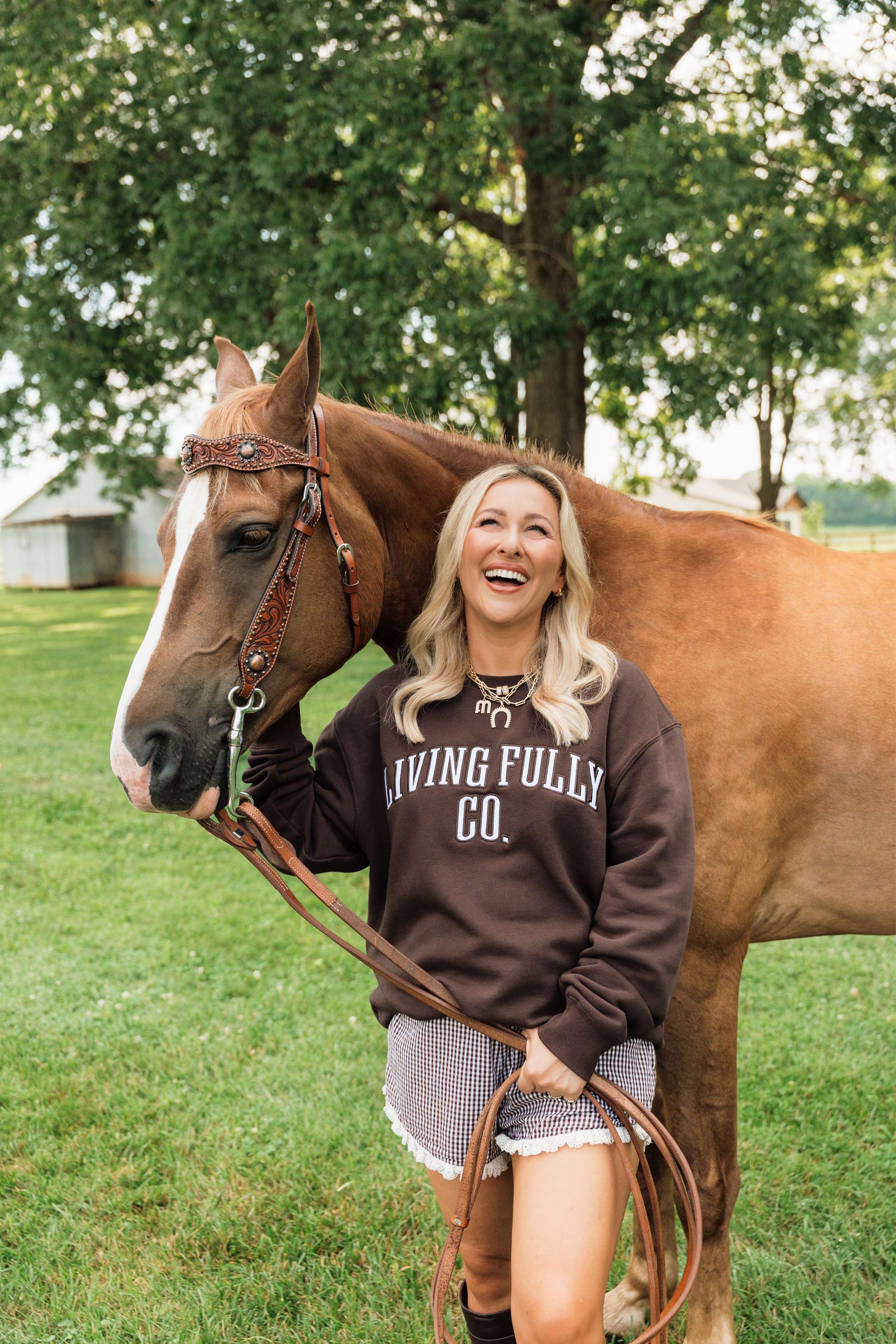 A woman smiling outdoors next to a brown horse, wearing a dark brown sweatshirt with 'LIVING FULLY CO.' printed in white across the chest.