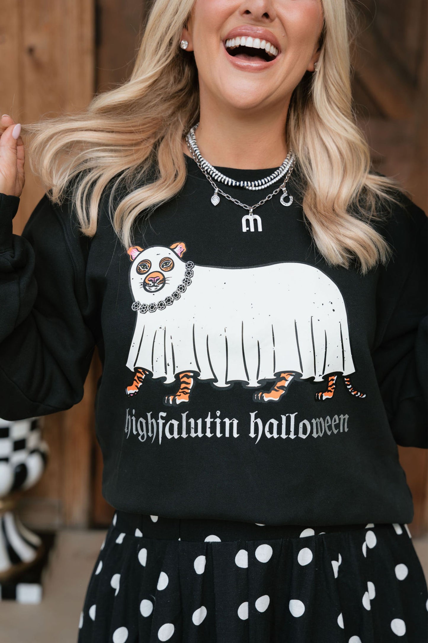 A woman wearing a black crewneck sweatshirt with a graphic of a white ghostly cat with orange paws and the text 'highfalutin halloween', paired with a black and white polka dot skirt.