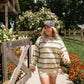 A woman walking outdoors on a garden path wearing a cream and olive green striped long-sleeve knit top, a brown baseball cap, white lace shorts, and beige knee-high boots.