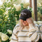 A woman wearing a black trucker hat with 'LIVING FULLY CO.' embroidered in white on the front, paired with a cream and olive striped sweater, posing outdoors in front of white hydrangea flowers.