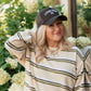 A woman wearing a cream and olive green striped knit sweater and a dark brown baseball cap with 'LIVING FULLY' embroidered on it, posing outdoors in front of hydrangea bushes.